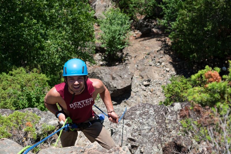 A man wearing a blue helmet and a maroon tank top with the word "Dexter" on it is rock climbing. He is holding a blue rope and is looking up with a smile. The background consists of rocks, trees, and other greenery. The man appears to be enjoying his climb.